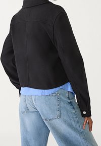 Osoba v zkrácené černé bundě přes světle modrou košili s světle modrými džíny, pózující s jednou rukou na bocích.