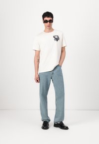 Vit kortärmad t-shirt med svart blommig grafik, i kombination med ljusblå raka jeans och svarta skor. Modellen bär solglasögon.