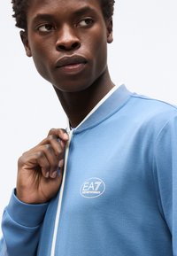 Νεαρός άνδρας τραβάει το φερμουάρ ενός ανοιχτόχρωμου μπλε μπουφάν Emporio Armani EA7 μπροστά σε λευκό φόντο.