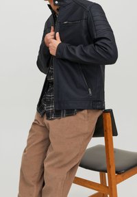 Giubbotto in pelle nera con zip asimmetrica, dettagli a coste sulle spalle e una vestibilità snella. Indossato sopra una camicia a quadri con pantaloni in corduroy beige.