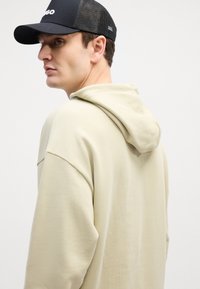 Felpa leggera beige con un fit rilassato e cappuccio integrato. Il tessuto appare morbido con una texture liscia. Presenta un berretto in rete nera.