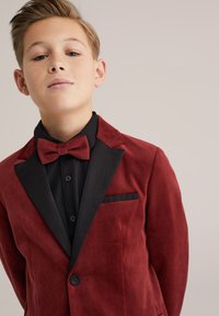 Raudonos aksominės medžiagos, pritaikytas švarkas su juodu satino atlapu ir kišene. Po juo juoda marškinėliai, papildytas derančia peteliške.