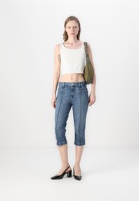 Top court blanc léger, pantalon capri en denim bleu avec poches avant et coutures contrastées, chaussures à talons blocs en vernis noir, sac olive.