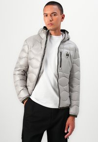 Grijze pufferjack met een geborduurd ontwerp, ritssluiting aan de voorkant, capuchon en een borstzak. Bevat zwarte en gele geribbelde accenten.