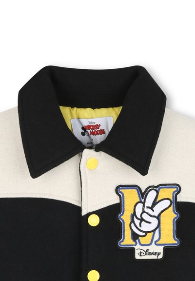 Giacca nera e crema con fodera interna gialla, caratterizzata da un grande "M" ricamato e un simbolo della mano. Bottoni gialli completano il design.