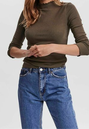 Femme portant un haut côtelé à manches longues vert olive et un jean bleu taille haute, les mains jointes devant, torse visible.