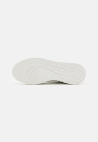 Suola di sneaker bianca in gomma testurizzata, con scanalature e un logo circolare. Il design include linee in rilievo per la presa e la stabilità.