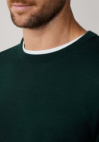 Uomo con barba che indossa un maglione a girocollo verde scuro sopra una maglietta bianca, mostrato dalla mascella al petto su uno sfondo neutro.