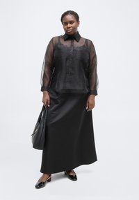 Γυναίκα που φοράει διάφανη μαύρη μπλούζα πάνω από ένα μαύρο τοπ, μακριά μαύρη φούστα, μαύρα γυαλιστερά παπούτσια με χαμηλό τακούνι και κρατά μια μαύρη τσάντα χειρός.