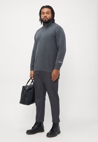 Grå genser med glidelås i halsen, ribbestrikket kant og ermer; kombinert med mørke bukser og svarte lærstøvler; holder en svart veske. Minimalistisk design.
