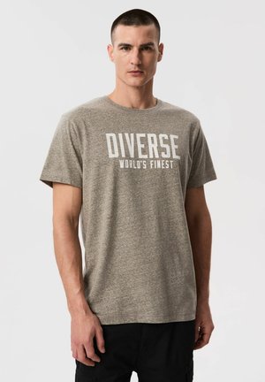 Mężczyzna o krótkich włosach, w szarej koszulce z napisem „DIVERSE WORLD'S FINEST”, stojący na jednolitym białym tle.