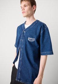 Denim kortermet skjorte i mørk blåfarge, med V-hals, knappen closures og en liten hvit logo på venstre brystlomme.