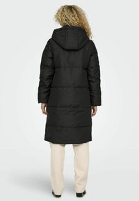 Longue doudoune noire avec capuche, présentant des coutures horizontales, une coupe ample et un ourlet légèrement évasé, portée sur un pantalon clair.