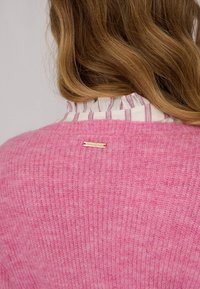 Pull en tricot rose avec une finition texturée, présentant un col rayé qui dépasse et un petit accent doré près du décolleté.