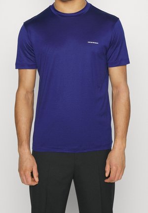 Mann trägt ein schlichtes dunkelblaues T-Shirt mit kurzen Ärmeln und kleinem "Emporio Armani"-Logo auf der linken Brust, kombiniert mit schwarzen Hosen.
