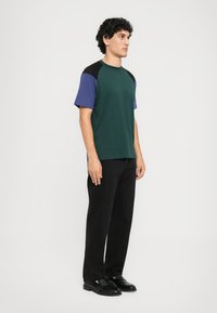 Uomo in piedi rivolto a destra, indossa una maglietta a maniche corte color-block, pantaloni neri e scarpe in pelle nere su sfondo semplice.