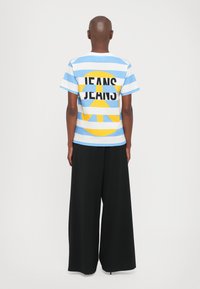 Stripet blå og hvit t-skjorte med gult sirkulært grafisk motiv og svart "JEANS" tekst på baksiden, kombinert med vide svarte bukser.