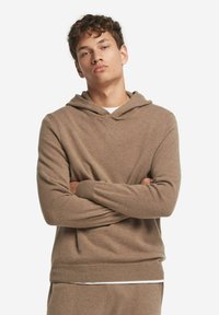 Junger Mann mit lockigem Haar, der einen braunen gestrickten Hoodie und passende Hosen trägt und mit verschränkten Armen vor einem weißen Hintergrund steht.
