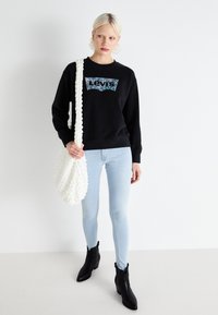 Čierna mikina s textúrovaným logom Levi's, svetlomodré úzke džínsy a čierne členkové topánky. Biela textúrovaná kabelka na rameno. Hladké textúry.