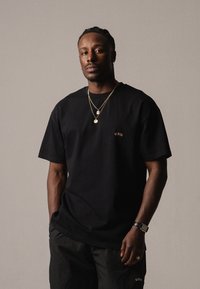 Camiseta de algodón negra con mangas cortas, que presenta un pequeño logo bordado en naranja. Combinada con pantalones cortos negros y cadenas de oro en capas.