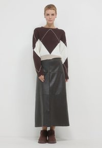 Donna in piedi contro un muro bianco indossa un maglione a rombi marrone e bianco, una gonna midi in pelle nera e stivali alla caviglia marroni.