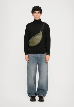 Mand iført sort turtleneck, brede falmede blå jeans, mørke sko og olivengrøn crossover-taske foran en ensfarvet hvid baggrund.
