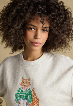 Young woman with curly hair wearing a light gray sweatshirt featuring an embroidered fox with a green jacket and beige pants.
