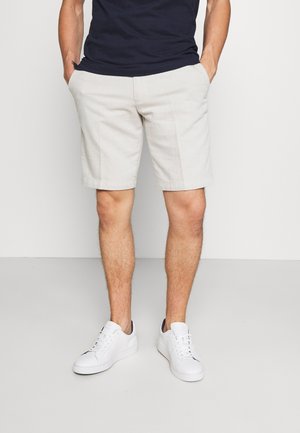 Pantaloni corti beige chiaro da uomo con taglio dritto, dotati di tasche laterali, abbinati a sneaker bianche e una maglietta blu navy.