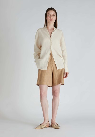 Camicia button-up in lino beige chiaro con colletto alla mandarina, abbinata a pantaloni corti in lino beige, e scarpe slip-on beige decorate con borchie.