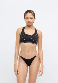 Μαύρο αθλητικό μπουστάκι με λευκό λογότυπο και σχέδιο, διαθέτοντας παχιά ζώνη. Μαύρο στρινγκ με λεπτή ζώνη μέσης. Ομαλό ύφασμα με ελαστικότητα.