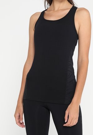 Frau trägt ein enganliegendes schwarzes ärmelloses Sporttop und passende schwarze Leggings, stehend vor einem schlichten weißen Hintergrund.