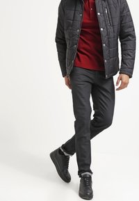 Giacca trapuntata nera sopra una polo rossa, abbinata a pantaloni slim-fit scuri e sneakers nere texturizzate. Design minimalista con dettagli funzionali.