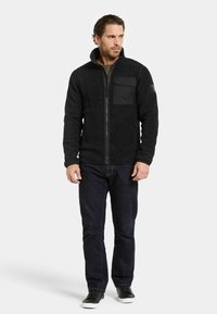 Zwarte fleecejack met een hoge kraag, een ritssluiting aan de voorkant en een contrasterend borstzakje; gecombineerd met donkere jeans en zwarte sneakers.