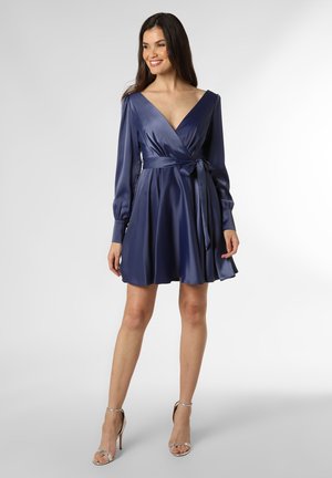 Navy satin wrap dress with long sleeves, v-neckline, and a tied waist detail, flowing into a flared skirt. Silver strappy heels accent the outfit.