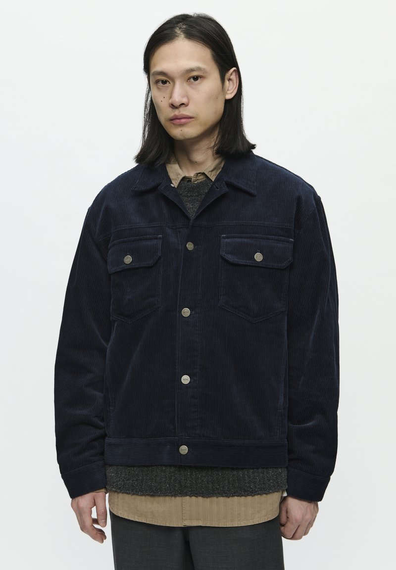 Námořnická kordová bunda s dvojicí náprsních kapes, zapínáním na knoflíky a mírně oversized střihem. Přes šedý svetr a béžovou košili.