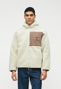 Jaqueta de fleece creme da Calvin Klein com capuz, apresentando um fecho frontal com zíper e um bolso castanho com detalhes do logótipo no lado esquerdo do peito.