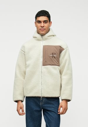 Calvin Klein cream fleece jacket with a hood, featuring a front zip closure and a brown pocket with logo detail on the left chest.