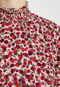 Blomstrig blus med volangkrage, prydd med röda, rosa och gröna blommor på en ljus krämfärgad bakgrund. Mjuk tyg med lätt struktur.