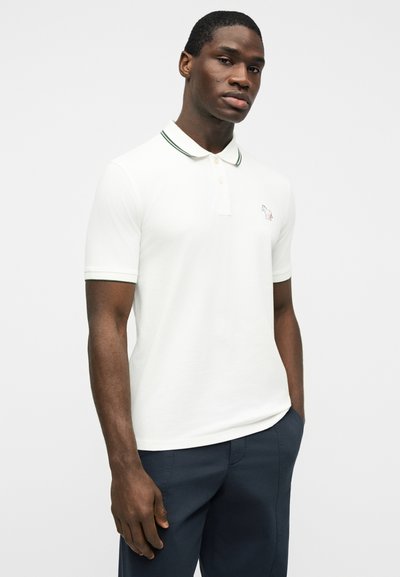 Άνδρας που φοράει λευκό πόλο πουκάμισο με γιακά και άκρα μανικιών με πράσινο περίγραμμα, σκούρο παντελόνι, στέκεται με το ένα χέρι στην τσέπη.