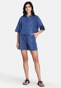 Kortärmad, croppad blå skjorta med krage, ihop med matchande blå shorts. Tillverkad av texturerat tyg. Buren med mörka sandaler.