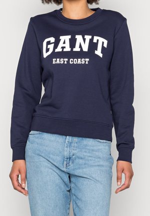 Sweat-shirt bleu marine avec inscription blanche "GANT EAST COAST". Manches longues, col rond, poignets et ourlet côtelés. Porté avec un jean bleu clair.