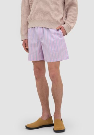 Person wearing pink and blue striped shorts, beige knit sweater, and tan slip-on shoes against a plain light background.