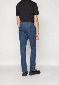 Pantalon bleu pour homme en coton, avec une coupe droite et des poches arrière ornées de boutons. Porté avec une chemise noire à manches courtes.