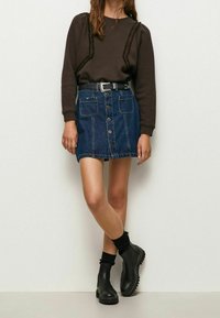 Sudadera marrón oscuro con detalles texturizados, combinada con una falda de mezclilla en línea A con botones frontales y dos bolsillos. Botines negros.