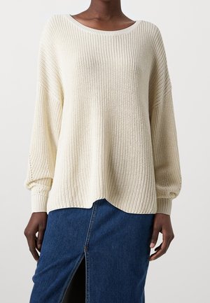 Persoon draagt een crèmekleurige geribbelde gebreide trui met lange mouwen en een ronde hals, gecombineerd met een blauwe denim rok met een split aan de voorkant.