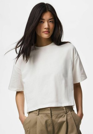 Jeune femme aux longs cheveux foncés, portant une chemise blanche ample à manches courtes et un pantalon beige taille haute, les mains dans les poches.