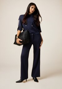 Completo blu navy con un top largo con cappuccio e accenti argentati, abbinato a pantaloni a gamba larga; borsa nera con hardware dorato.