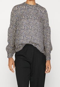 Blouse à manches longues avec motif floral bleu et beige associée à un pantalon taille haute noir porté par une personne debout.