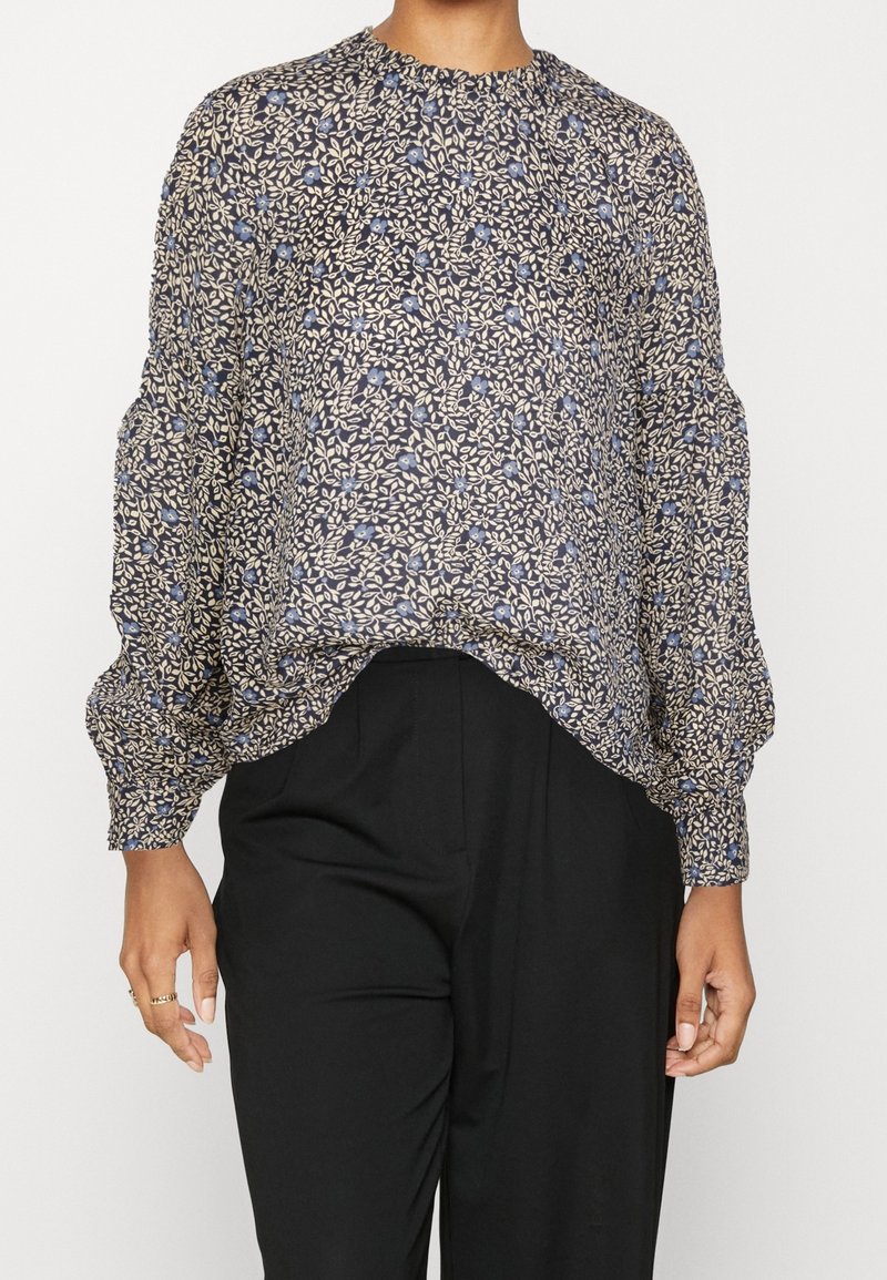 Blouse à manches longues avec motif floral bleu et beige associée à un pantalon taille haute noir porté par une personne debout.