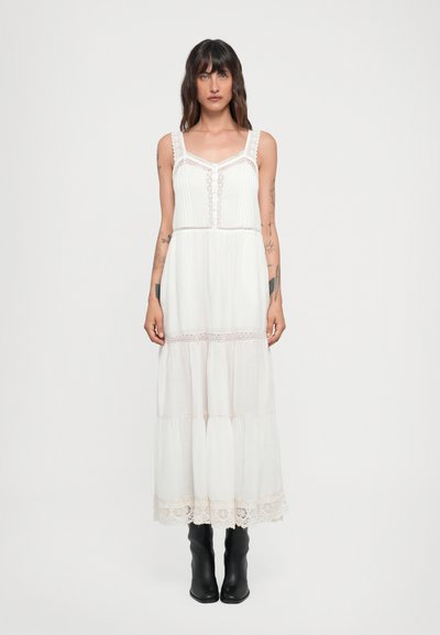 White maxi dress with lace accents, pleated bodice, and tiered skirt. Features adjustable straps and a delicate lace hem. Worn with black boots.